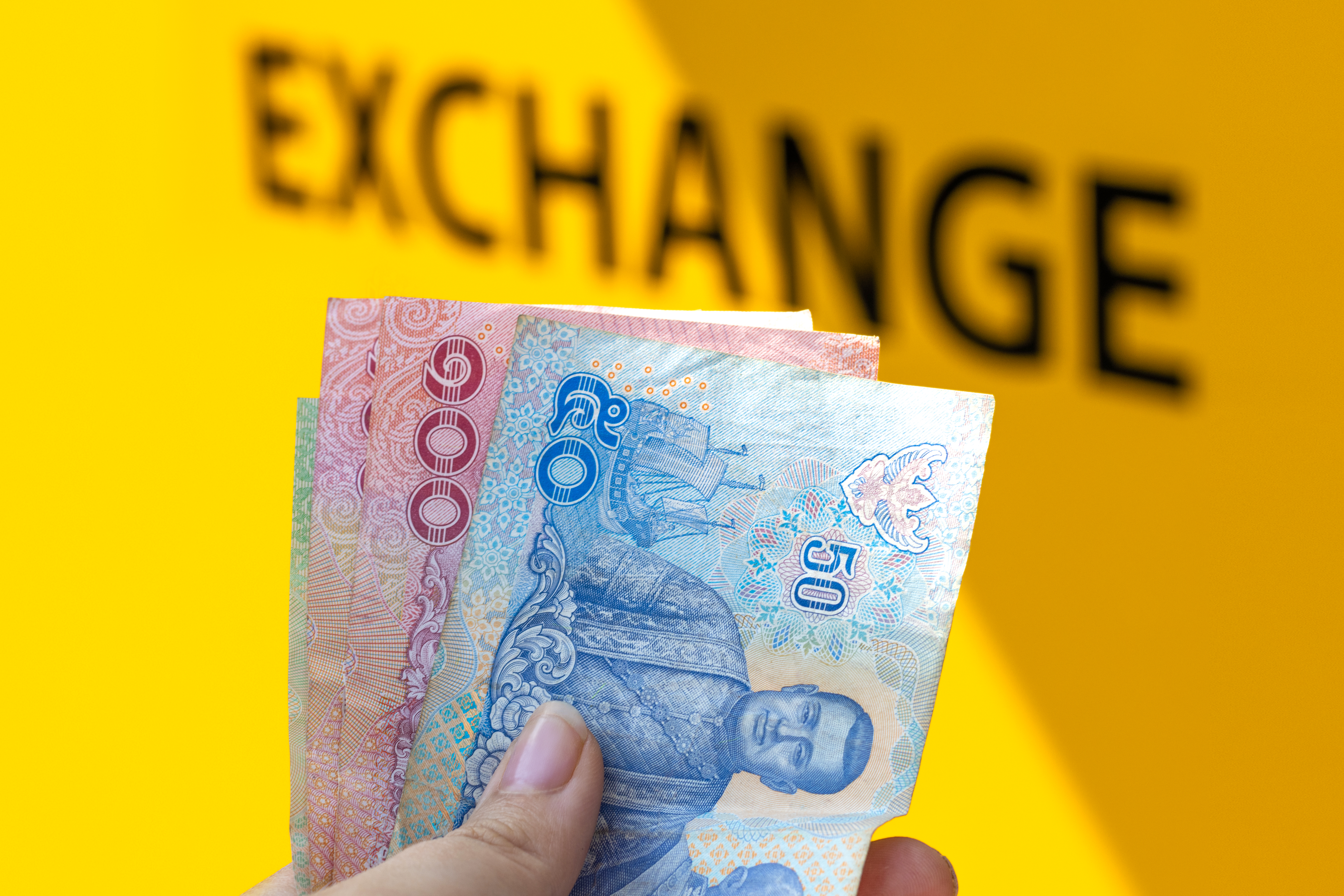 A hand holds a stack of Thai Baht banknotes - 50 and 100 Baht - against a blurred yellow background with the word 'EXCHANGE' visible. This image represents currency exchange and the Thai economy.